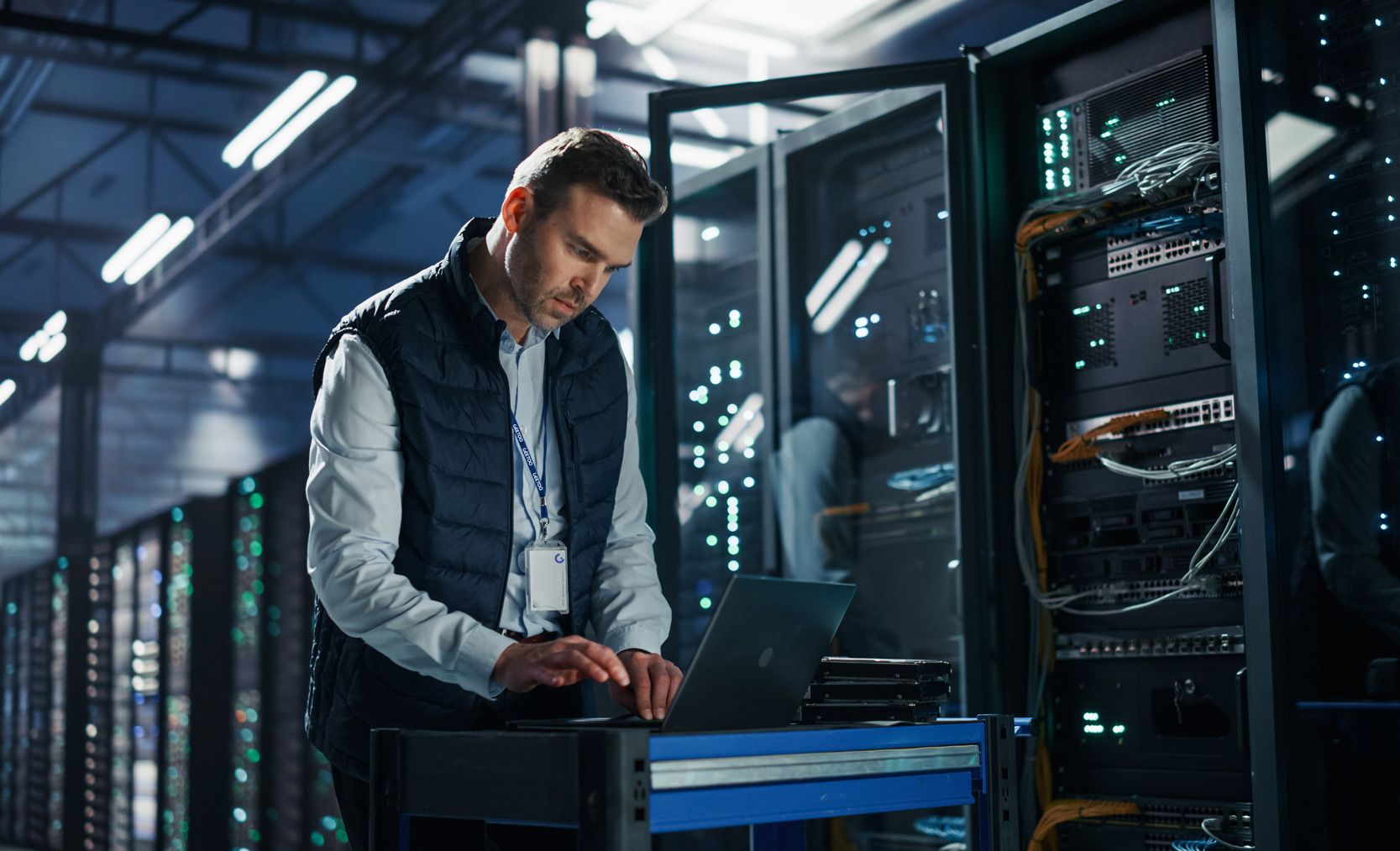IT technician in a server room typing on a laptop next to a server rack.